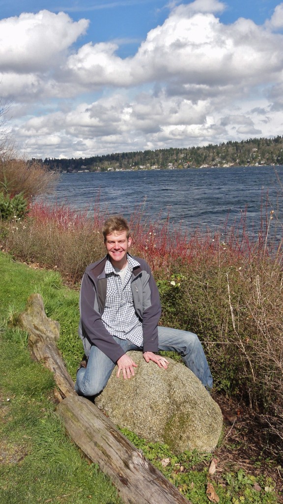 Image shows a young man sitting astride a granitic boulder, with Lake Washington and Mercer Island in the background.