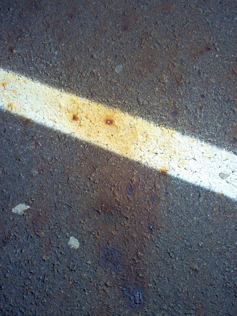 Image shows a parking lot stripe and a bit of the asphalt around it. Both the asphalt and the white stripe are spotted with rust-colored scorch marks. It appears that rain or snowmelt have smeared out the marks somewhat, but you can see the dots were the hot cinders landed.