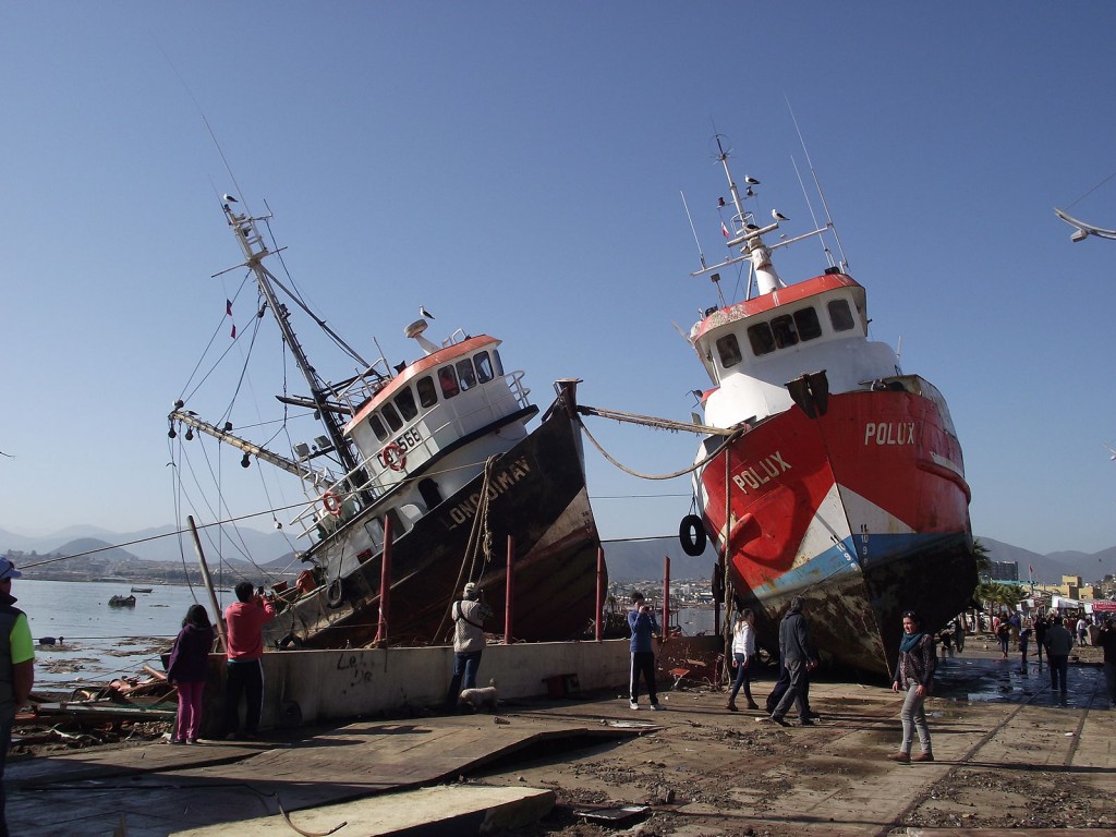 Image shows two large fishing boats stranded on the shore. Both of them are tilting at drunken angles. The pavement they're resting on has been buckled by the quake. People are milling around, and seagulls are perched in the rigging.