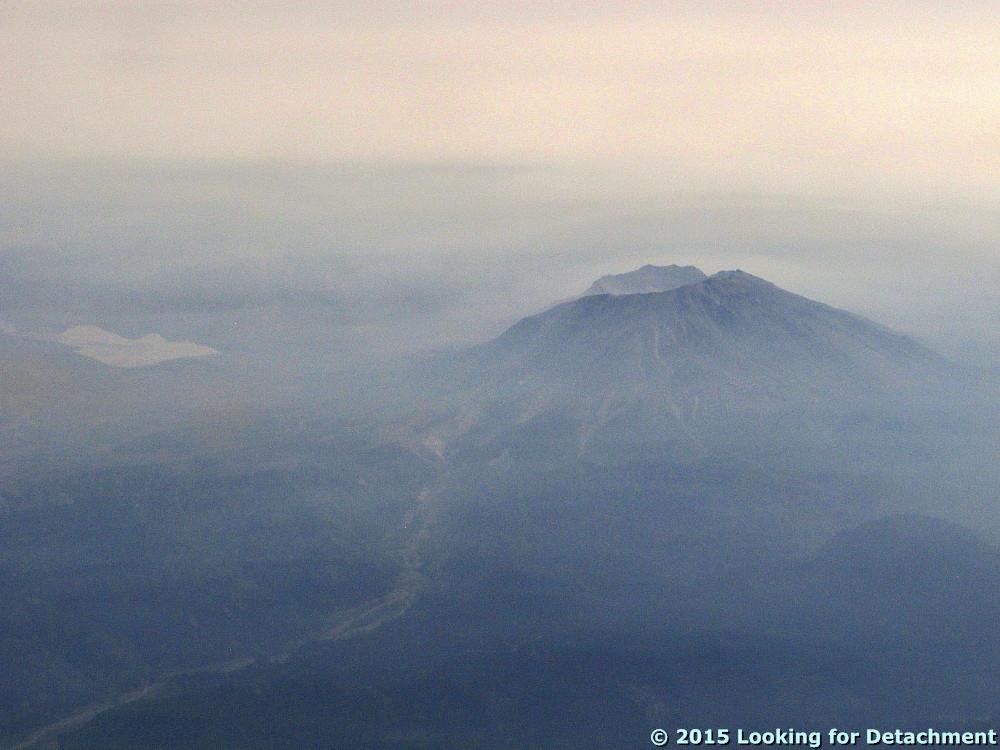 Image shows Mount St. Helens from 30,000 or so feet. The whole image looks like the aftermath of a volcanic eruption, everything basically varying shades of purple-gray. Mount St. Helens is a darker lump with a sliced-off top. You can see one of the forks of the Toutle River and Spirit Lake. The horizon vanishes in a haze.