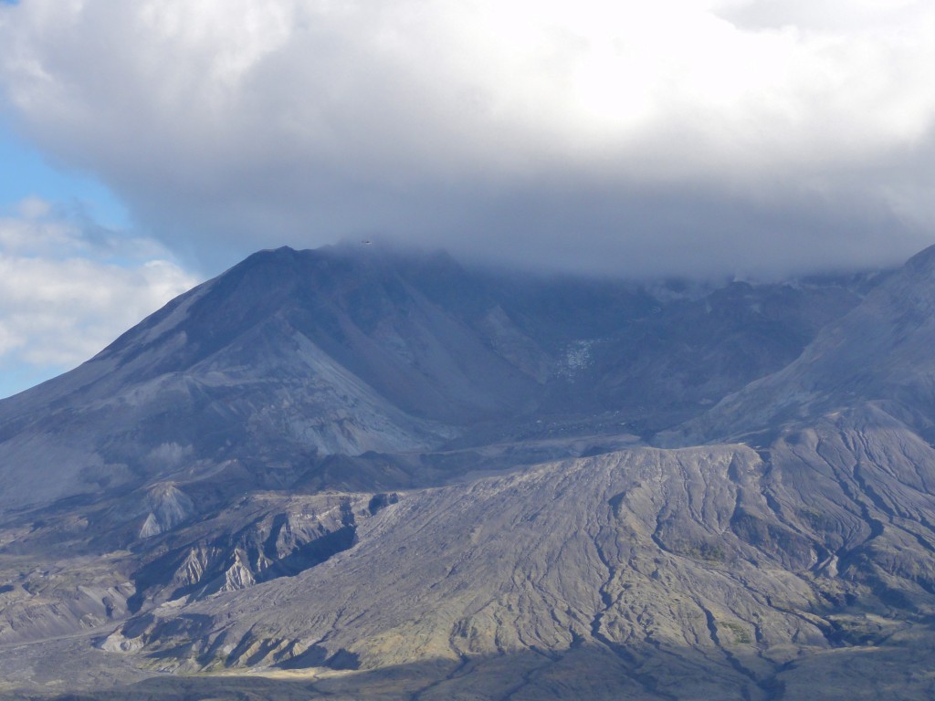 Image shows a portion of Mount St. Helens. The upper rim of the crater is obscured by cloud, but the interior of the crater, exterior wall, and ramp of pyroclastic deposits spilling from the gap in the rim are all visible. The dome is a low, wide mound within. The helicopter is flying past the rim. It's only a few pixels wide and virtually invisible.