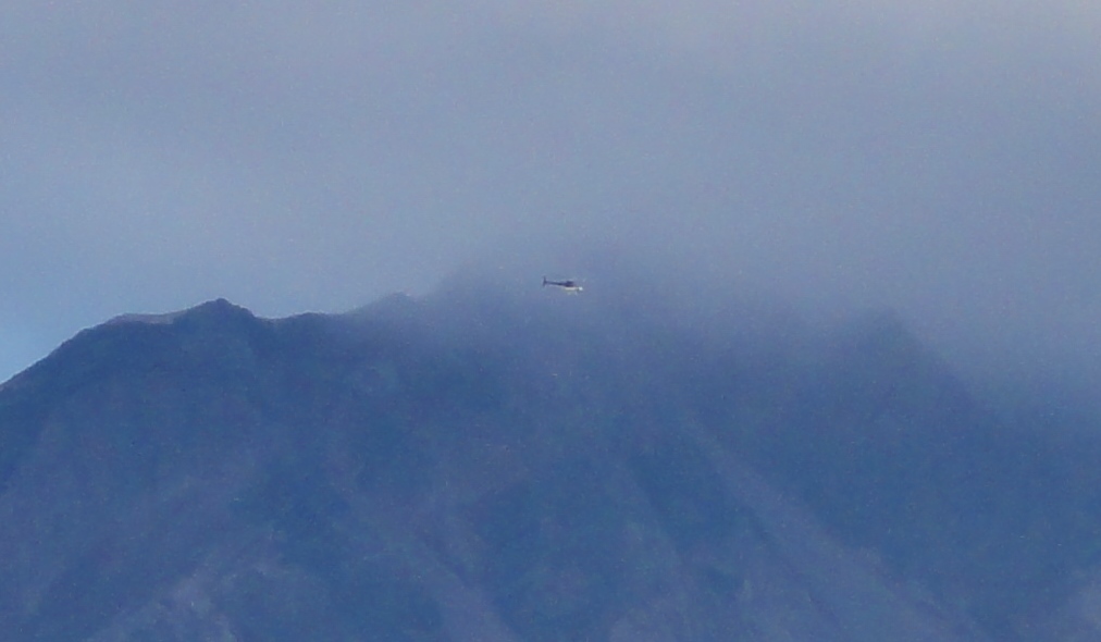 Image is a crop of the previous, showing a piece of the crater rim and the overlying cloud. The helicopter is just visible as a tiny streak between the cloud and the crater.