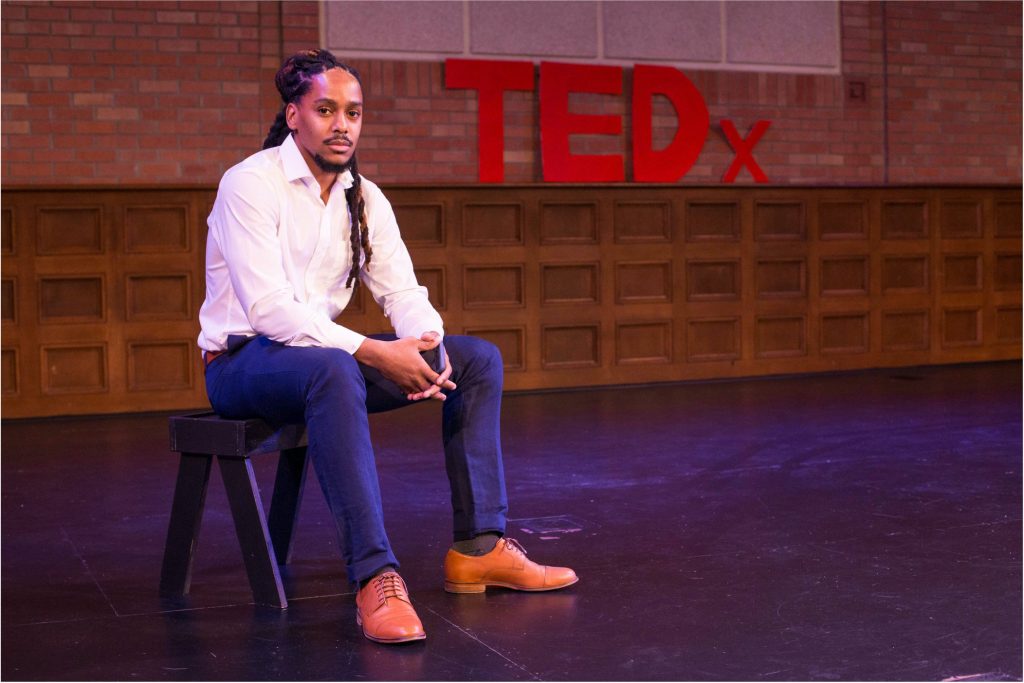 Photo of Justin Hardiman, wearing white dress shirt, jeans, and tan leather lace-up shoes, seated, against a dark backdrop with large red lettering: "TEDx"