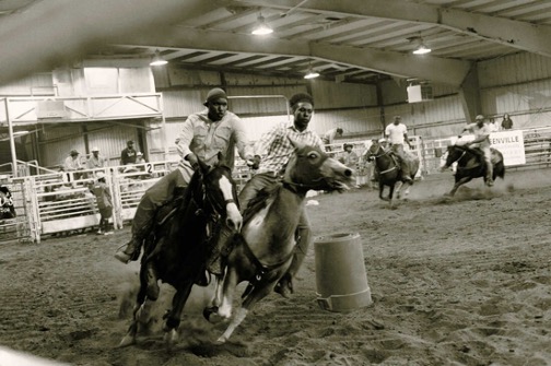 Black & white photo of four Black men on horses racing around a course in an indoor arena.