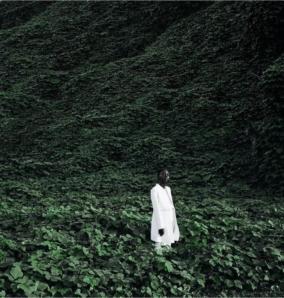 A Black model in a white mid-length coat stands in, and in front of, a field and backdrop of thick, dark green vines.