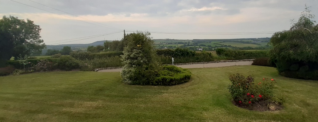 This photo shows the garden in which I'm writing (the clump of bushes by the right edge of the photo seems to have a nest of baby birds in it, who make a racket every few minutes), and a big ol' hill across the river valley. The hill is mostly pasture land, dotted with a few cows and sheep, but there are trees between the pastures, and what looks like a forest of very evenly-sized pine trees along the ridge, which makes me wonder if they're intended to be lumber. To the left, near the edge of the ridge, you can see a lone wind turbine. The clouds overhead are a little patchy, letting through glimpses of the evening sky and its colors. 